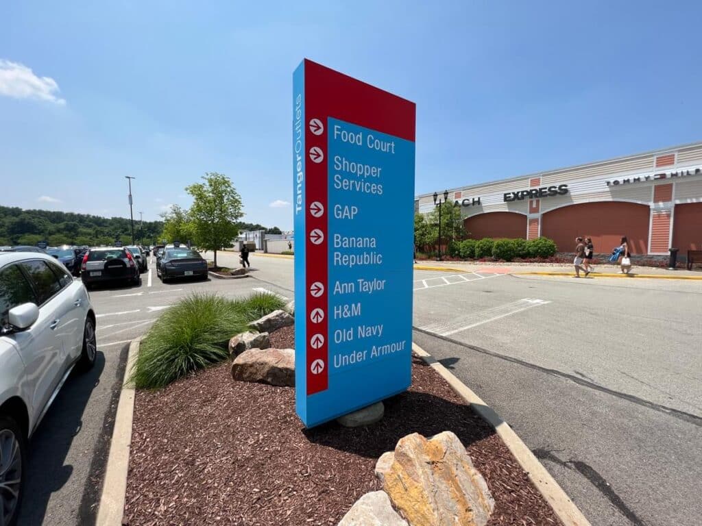 Tanger Outlets wayfinding pylon sign near parking area with shopper services and store directory.