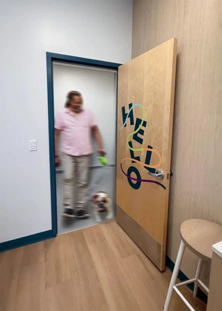 A person walking a small dog enters a veterinary exam room through a wood door decorated with colorful vinyl graphics and the word “HELLO” arranged vertically.